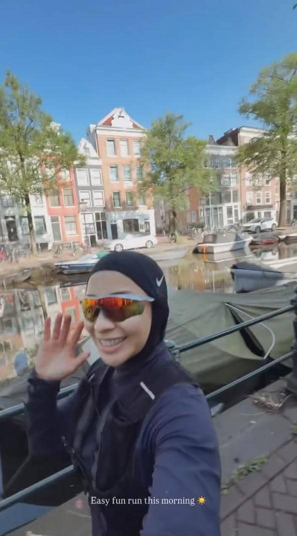 Seorang perempuan mengenakan pakaian olahraga dan kacamata hitam berpose di tepi kanal dengan latar bangunan khas Belanda di bawah langit cerah.