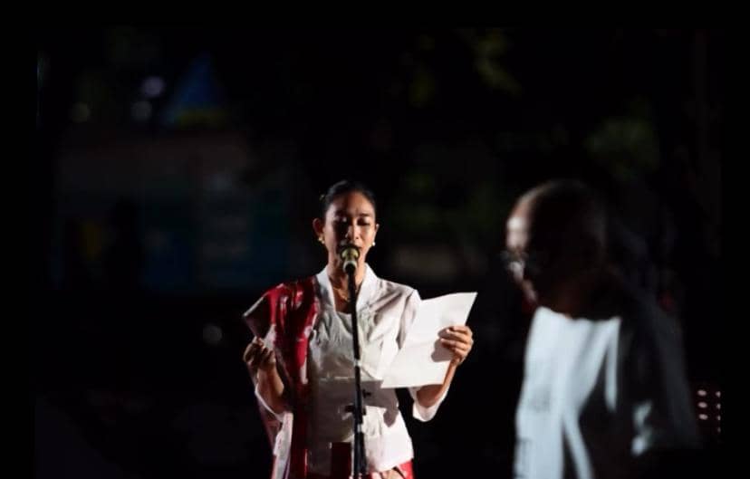 Seorang perempuan mengenakan pakaian putih dan selendang merah sedang membaca doa di depan mikrofon pada acara doa bersama malam hari.