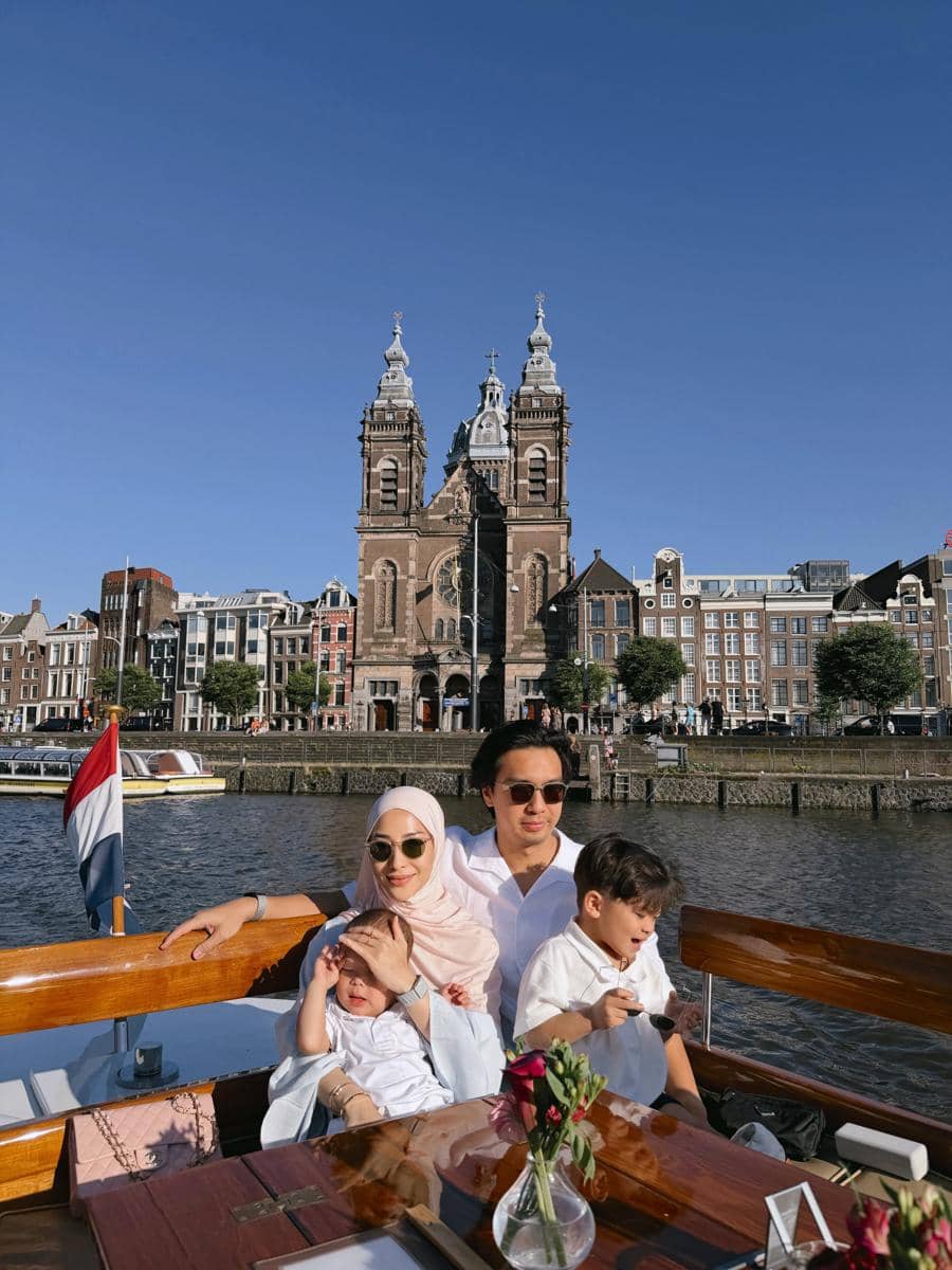 Sebuah keluarga menikmati perjalanan perahu di kanal Amsterdam dengan latar belakang Gereja Santo Nikolas di bawah langit biru cerah.