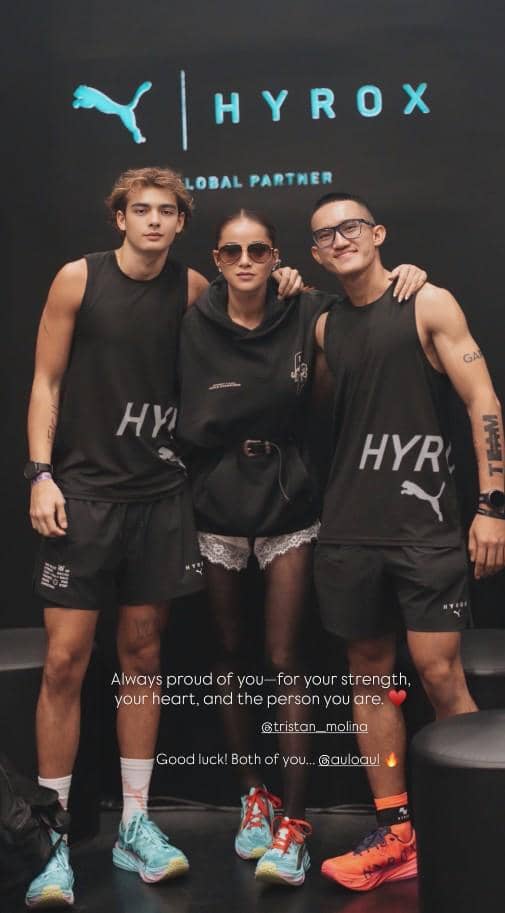 Tiga orang berpose di depan latar bertuliskan HYROX dan logo Puma, mengenakan pakaian olahraga hitam dengan sepatu berwarna cerah.