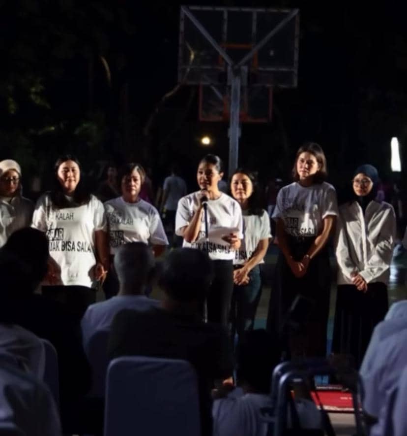 Sekelompok orang mengenakan kaus putih berdiri di depan audiens saat acara doa bersama pada malam hari di area terbuka.