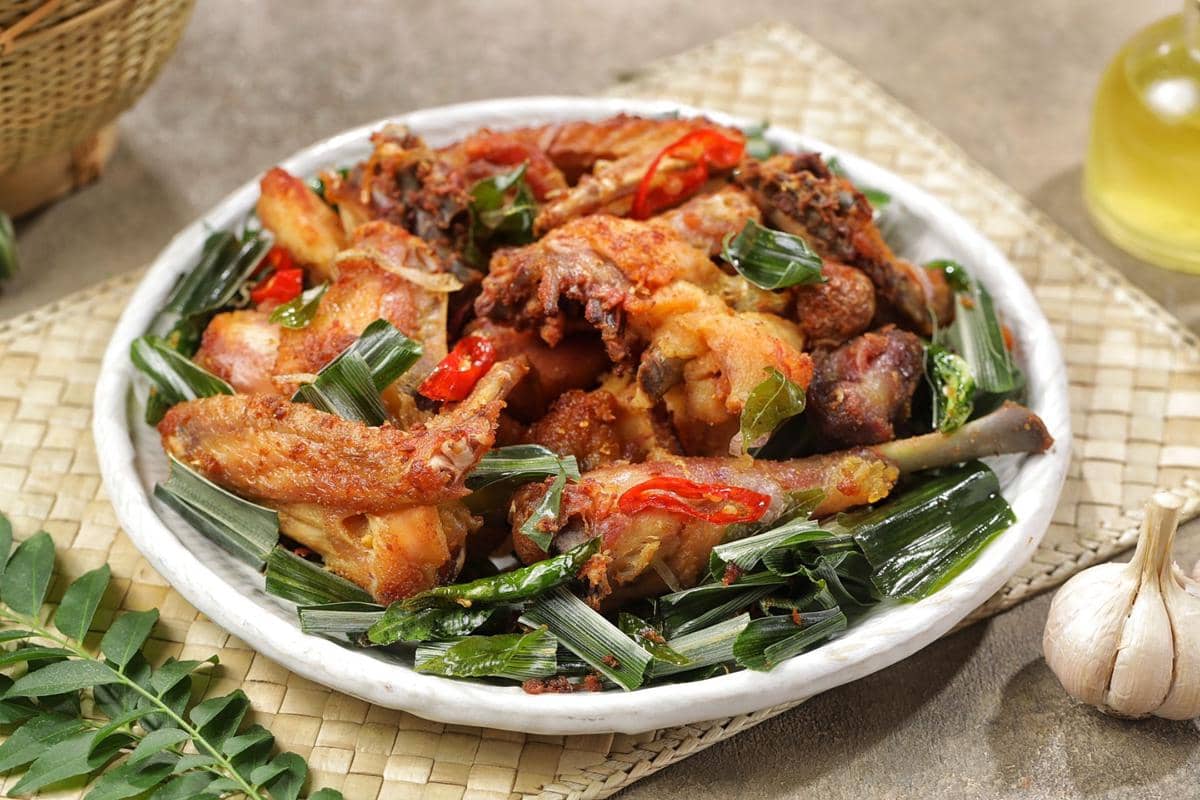 Sepiring ayam goreng kalasan berwarna keemasan disajikan dengan irisan cabai merah dan daun pandan di atas anyaman bambu.