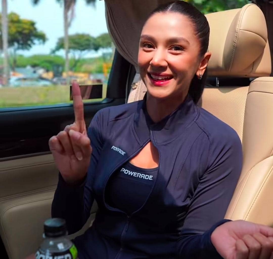 Seorang perempuan mengenakan jaket olahraga biru tua duduk di dalam mobil sambil tersenyum dan mengangkat satu jari tangan.