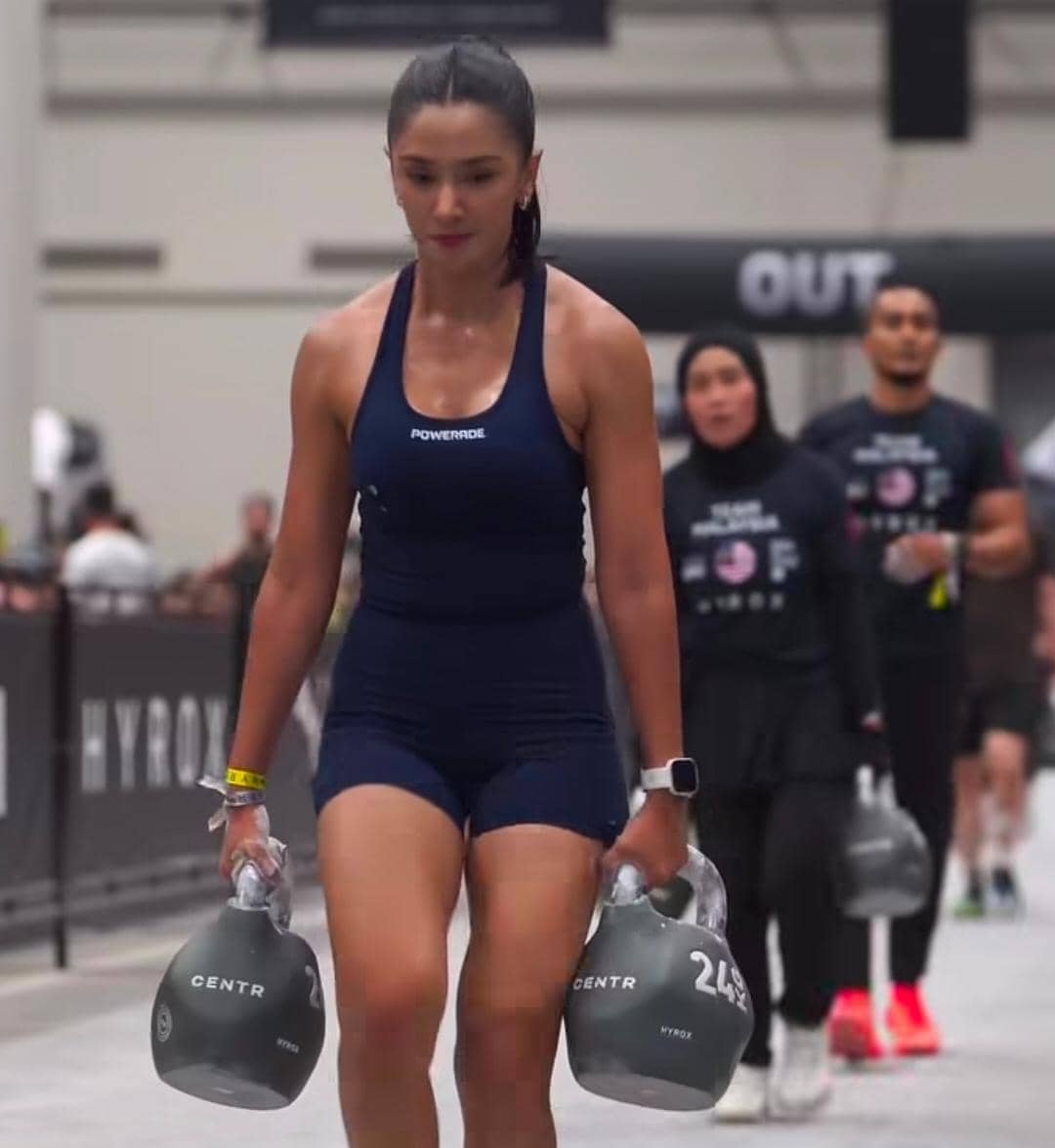 Seorang wanita mengenakan pakaian olahraga biru tua sedang membawa dua kettlebell di arena kompetisi Hyrox indoor.