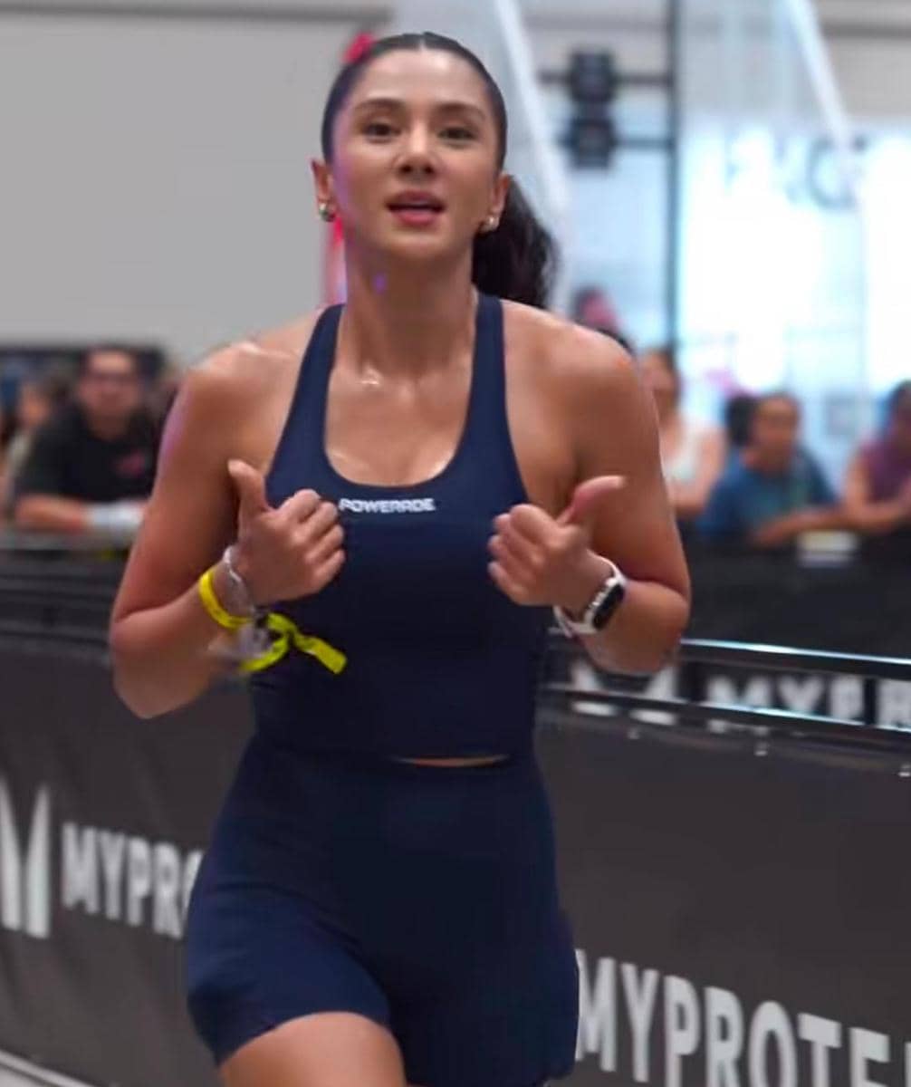 Seorang perempuan mengenakan pakaian olahraga biru tua sedang berlari di arena kompetisi kebugaran indoor dengan ekspresi semangat.