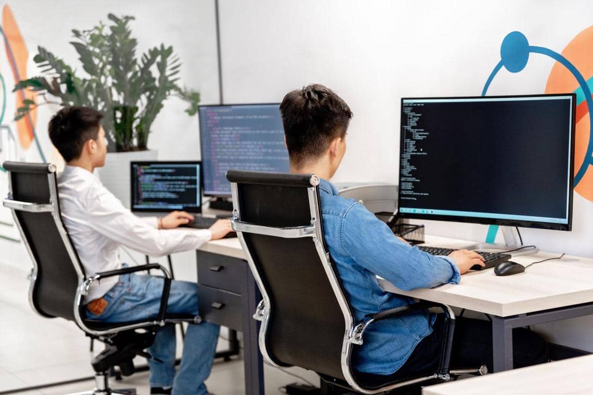 ilustrasi dua pria sedang duduk di meja kerja dan melakukan pemrograman di ruang kerja kantor modern dengan monitor besar