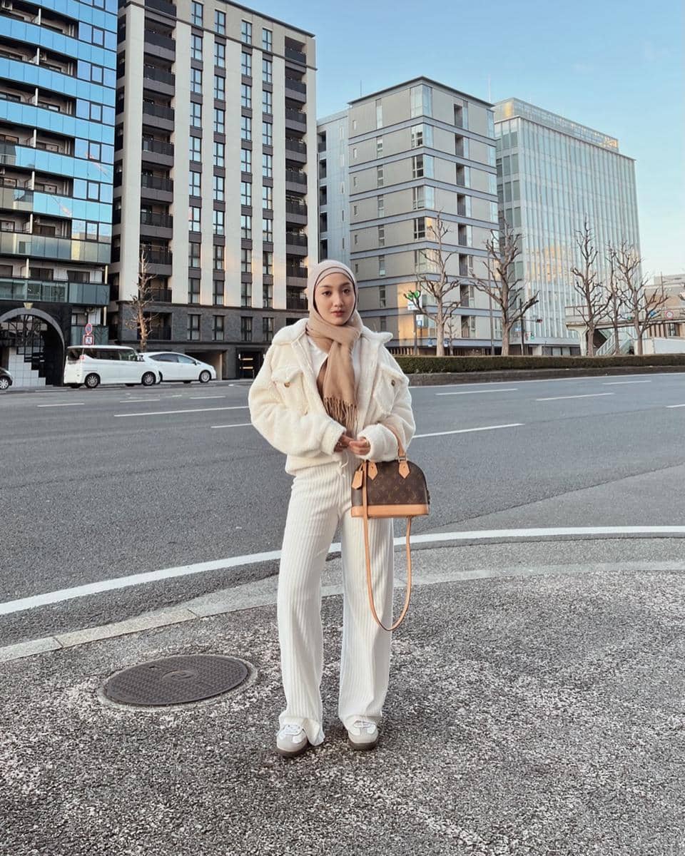 Seorang wanita berhijab mengenakan outfit musim dingin berwarna krem dengan jaket bulu dan celana panjang berdiri di tepi jalan kota modern.