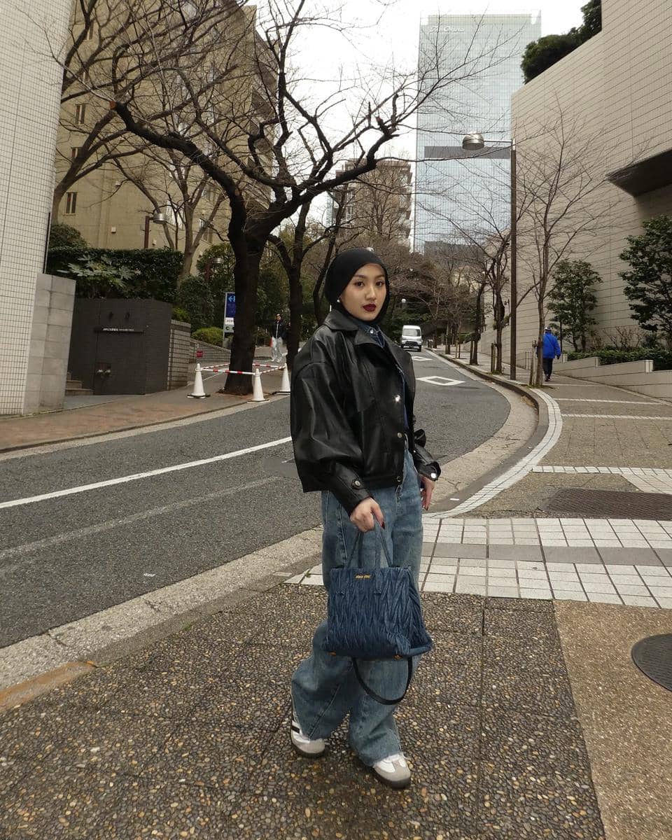 Seorang wanita berhijab mengenakan jaket kulit hitam, celana jeans longgar, dan tas biru berpose di jalan kota dengan latar gedung tinggi.