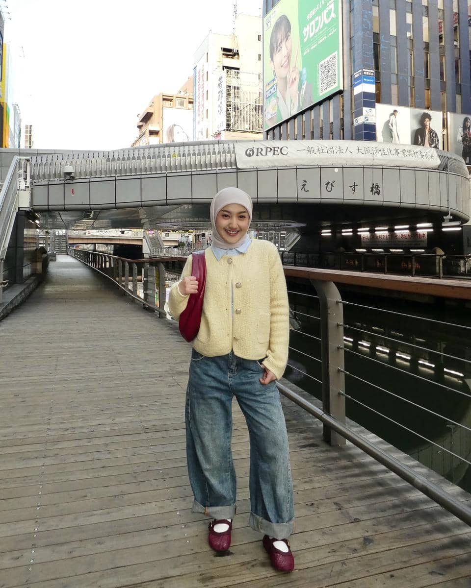 Seorang perempuan berhijab mengenakan cardigan krem, celana jeans longgar, dan sepatu merah berpose di jembatan kota dengan latar gedung bertulisan Jepang.