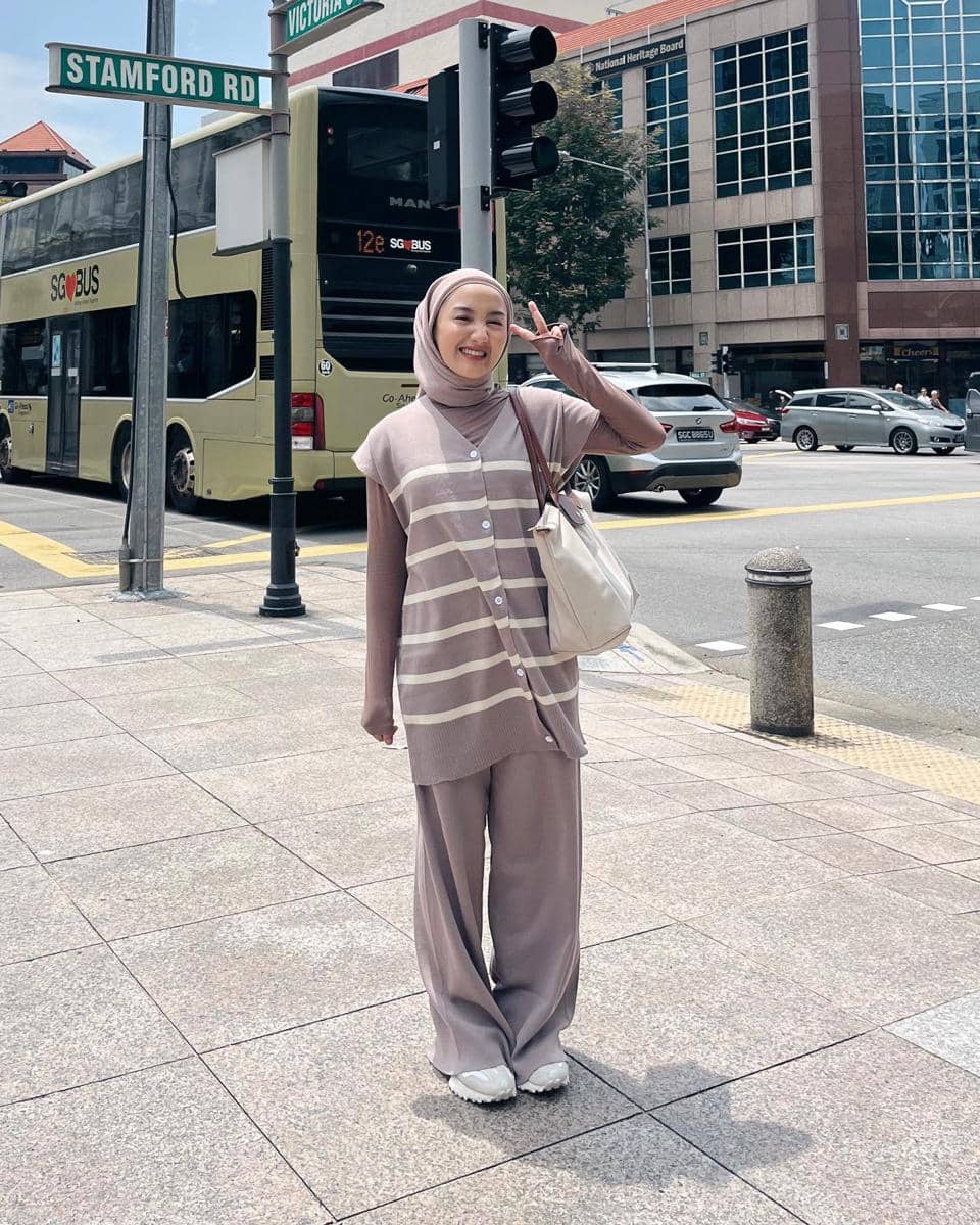 Seorang wanita berhijab berpose di persimpangan jalan Stamford Road dengan latar bus bertingkat dan gedung perkantoran modern.