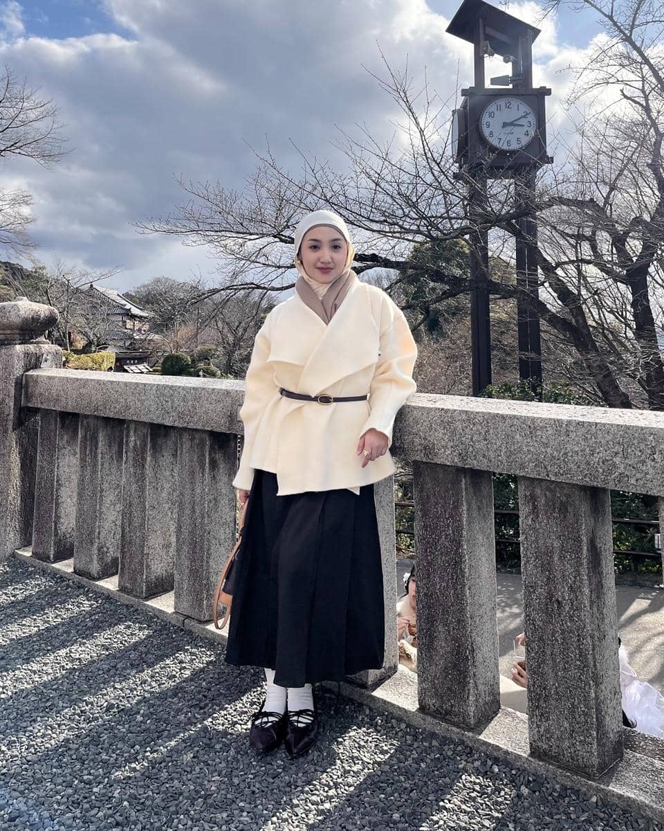 Seorang wanita berhijab mengenakan mantel krem dan rok hitam berpose di jembatan batu dengan latar jam menara dan langit berawan.