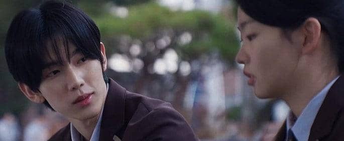 Dua siswa berseragam sekolah duduk berdampingan, salah satunya menatap temannya dengan ekspresi serius di adegan drama Agent Kim Reactivated.