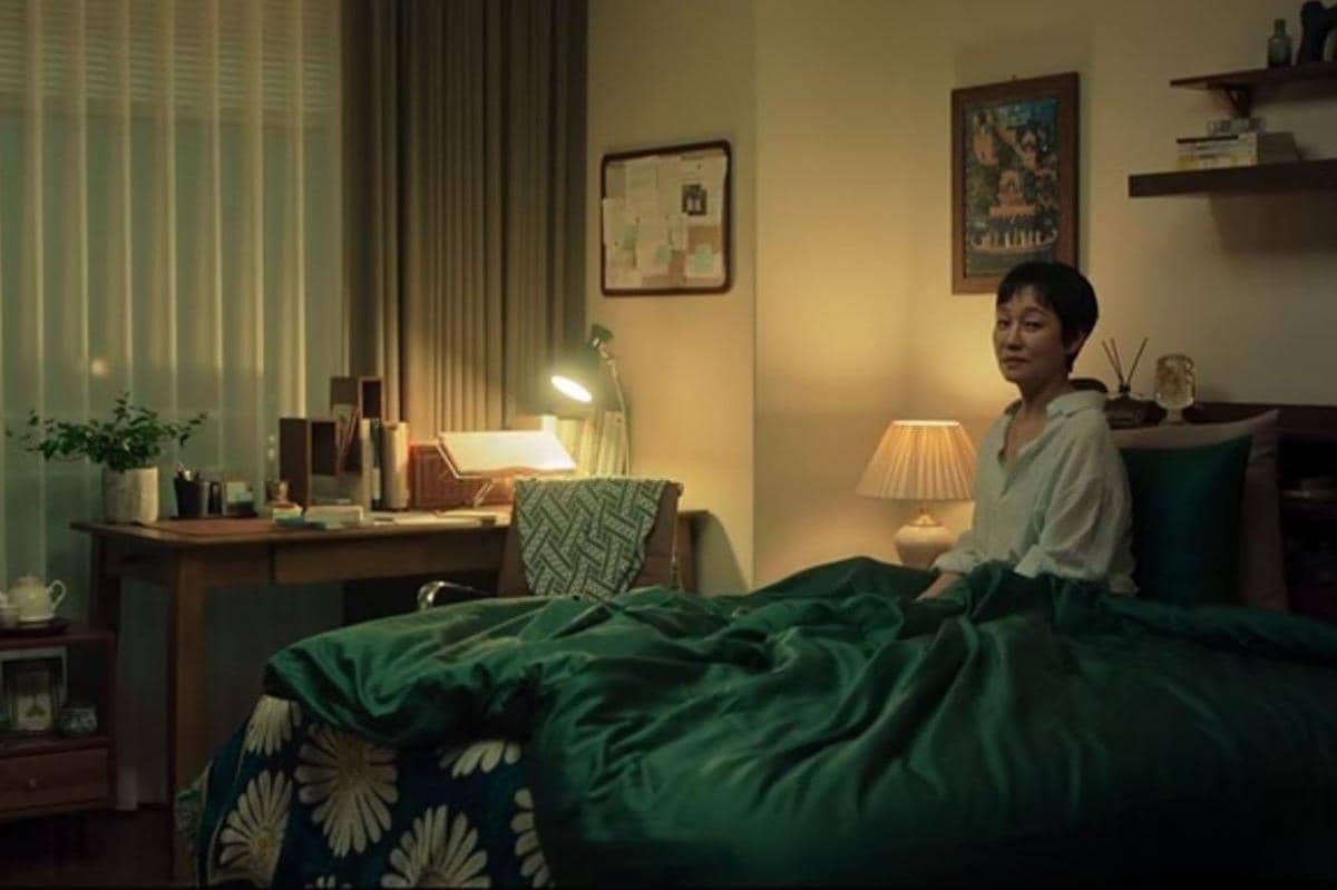 Seorang perempuan duduk di tepi tempat tidur dengan selimut hijau di kamar yang hangat dan lampu meja menyala lembut.