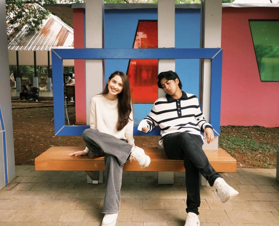 Dua orang duduk di bangku taman dengan latar warna-warni, satu tersenyum ke kamera sementara yang lain bersandar santai.