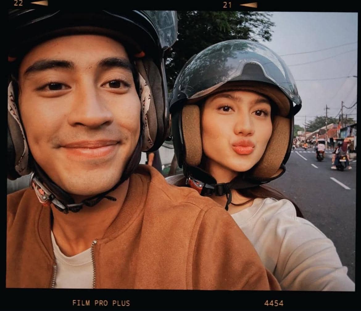 Sepasang pria dan wanita mengenakan helm sedang berpose di atas motor di jalanan kota saat sore hari dengan ekspresi bahagia.