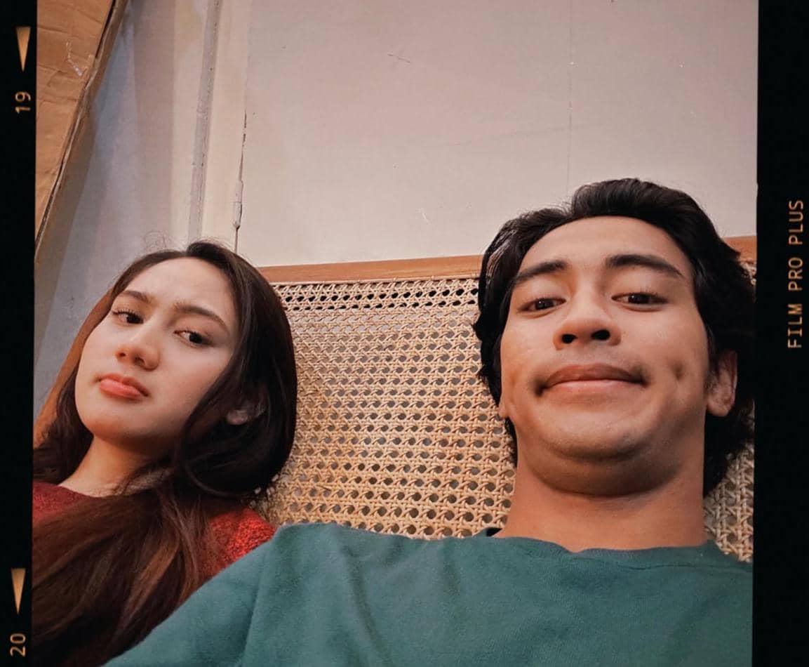 Dua orang duduk bersebelahan di kursi rotan, satu mengenakan baju merah dan satu lagi mengenakan baju hijau, berpose santai untuk swafoto.