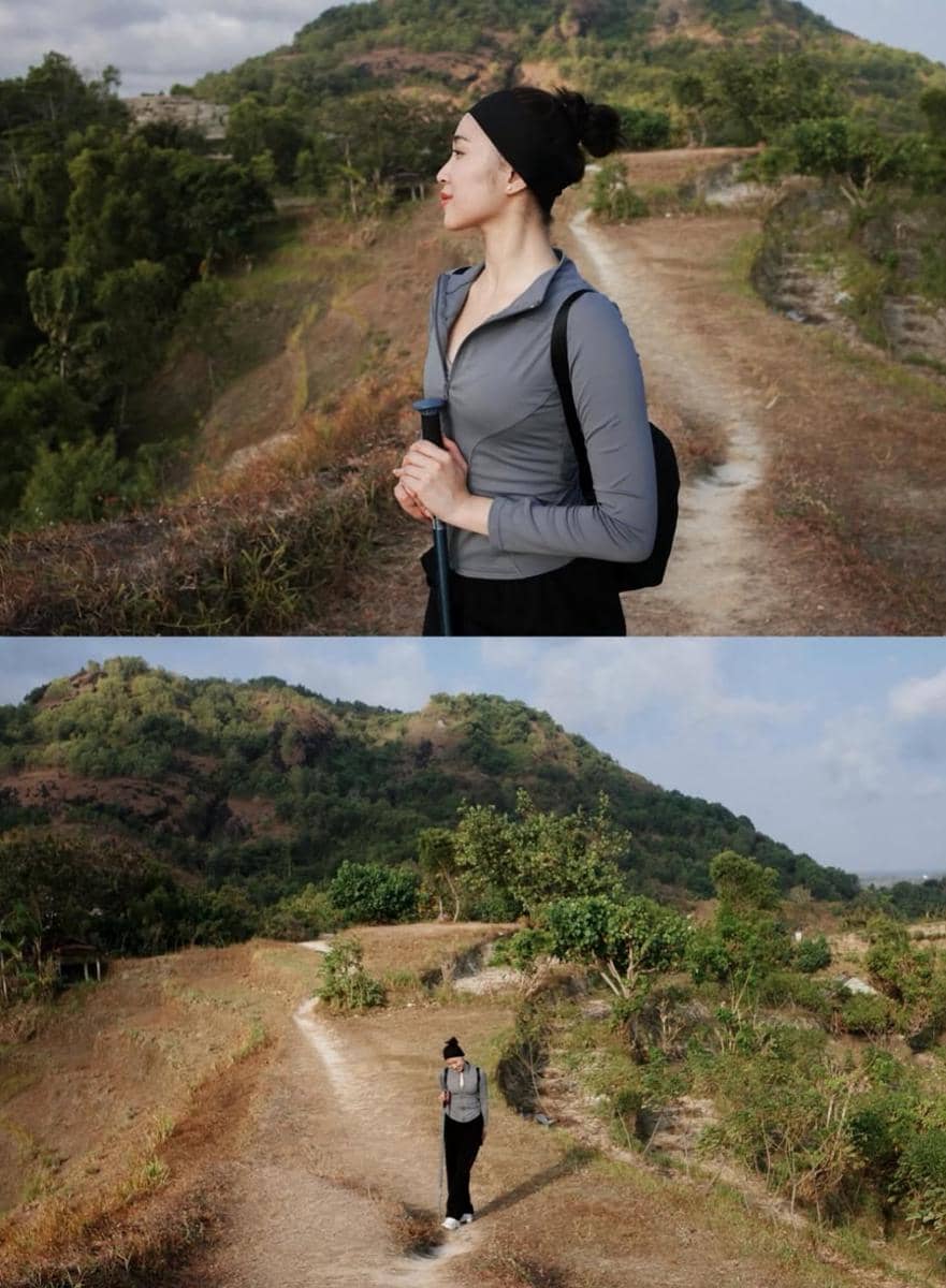 Seorang perempuan mengenakan jaket abu-abu dan ikat kepala hitam sedang hiking di jalur tanah dengan latar perbukitan hijau.