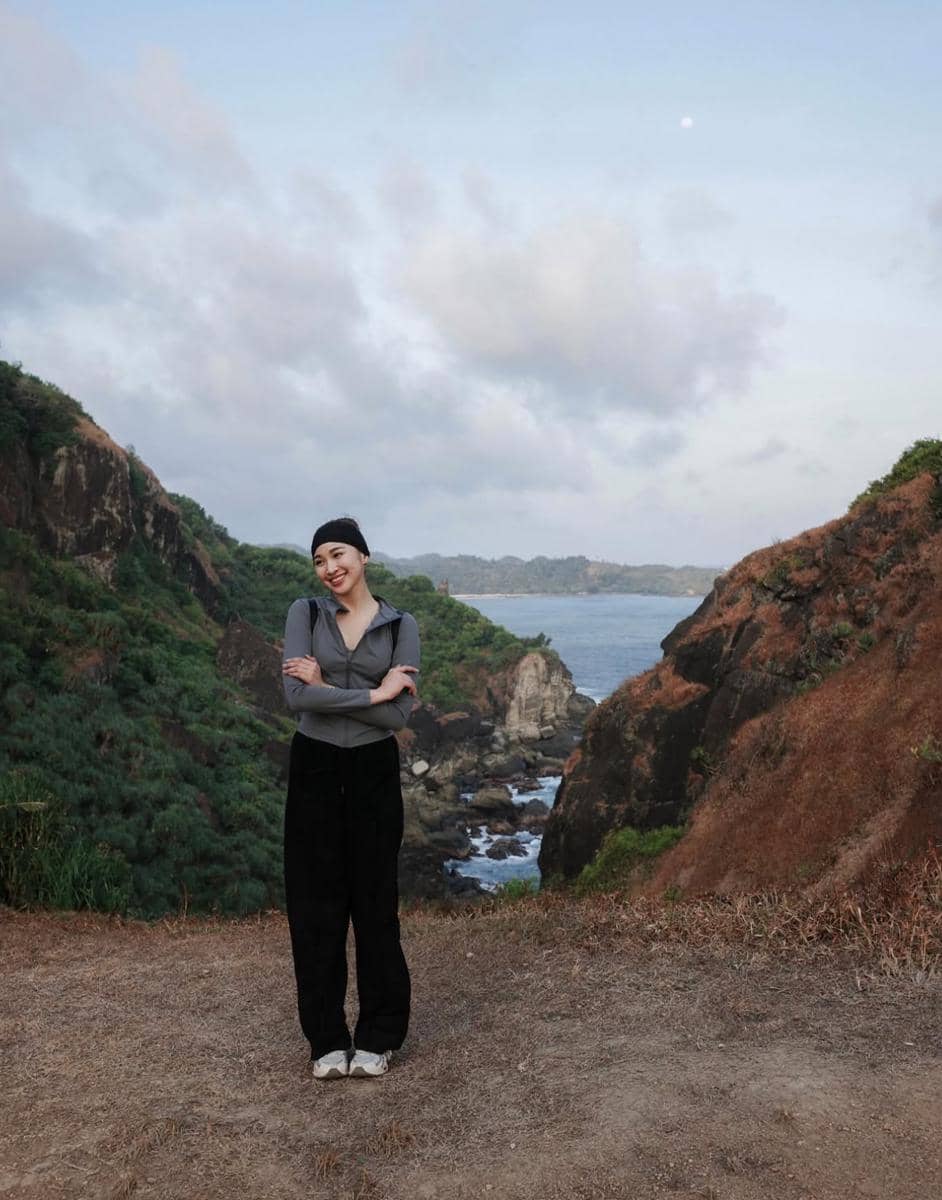 Seorang perempuan berdiri tersenyum di tepi tebing dengan latar laut dan perbukitan hijau kecokelatan di bawah langit berawan.