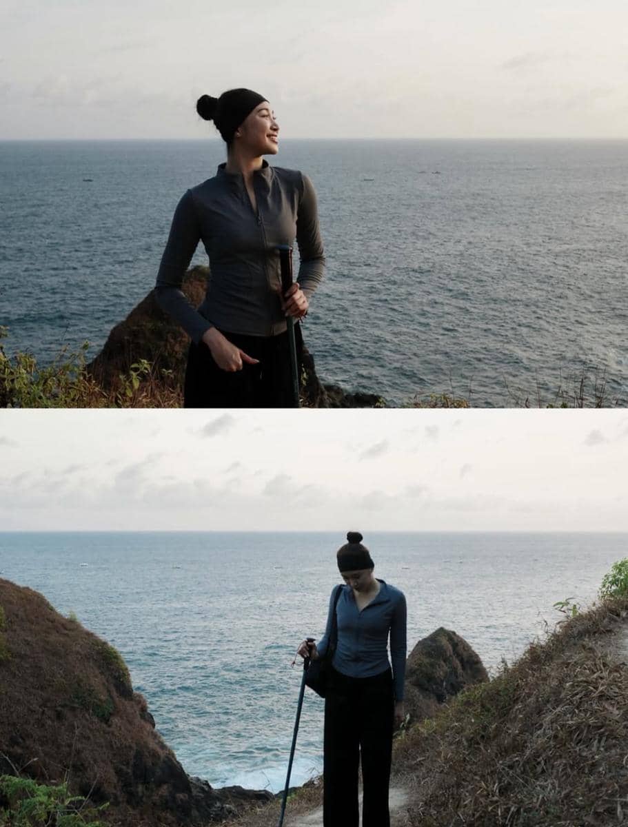 Seorang perempuan mengenakan jaket abu-abu dan topi hitam sedang hiking di tebing dekat laut dengan pemandangan samudra luas di belakangnya.