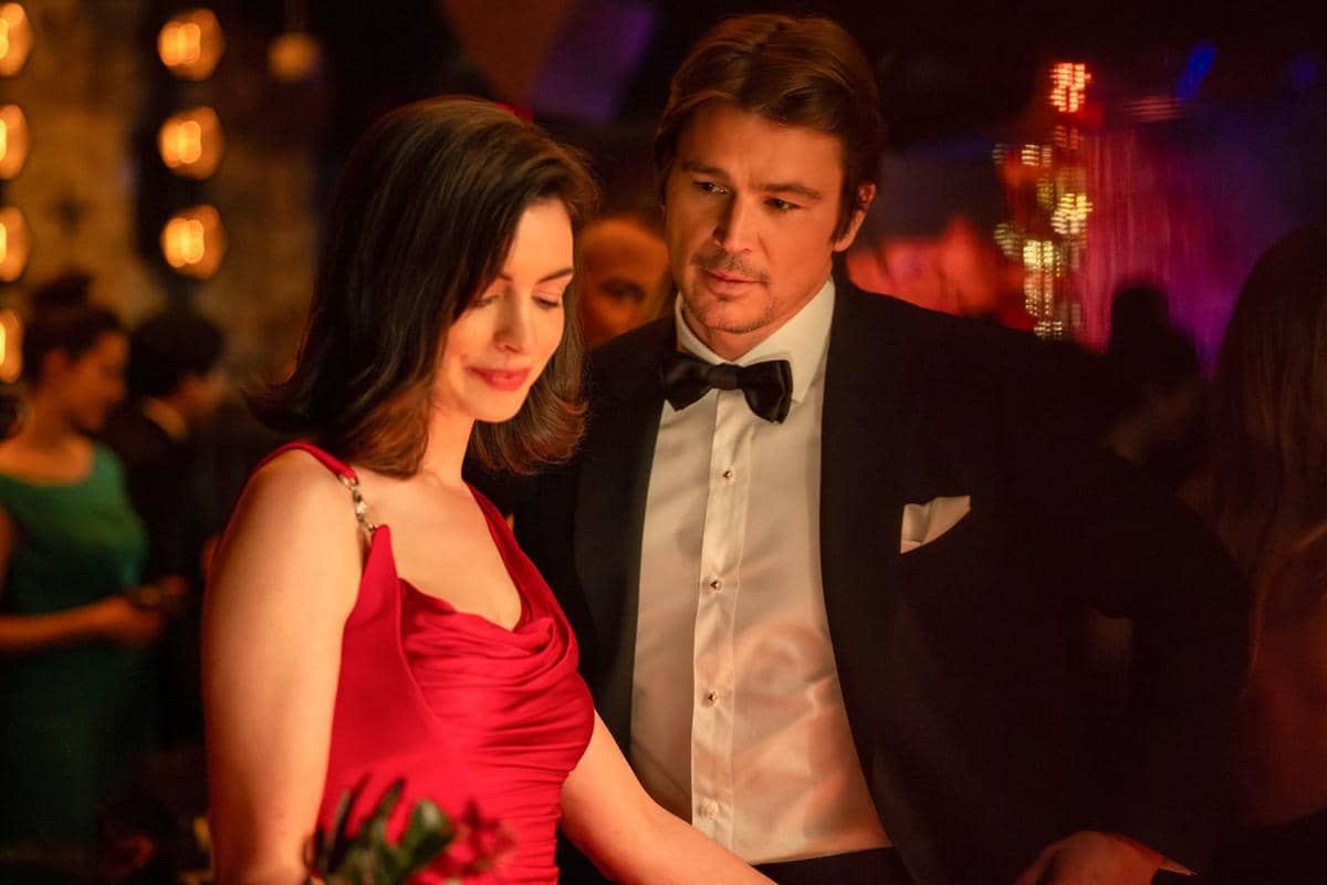 Dua karakter dalam film Verity berdiri berdekatan di pesta malam, wanita mengenakan gaun merah dan pria mengenakan setelan jas hitam.