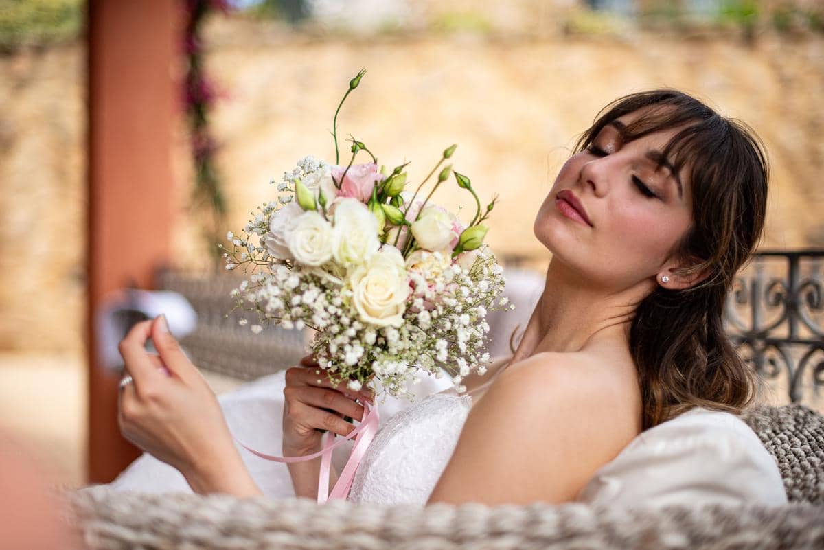 Seorang pengantin wanita berponi memejamkan mata dengan ekspresi wajah rileks sambil memegang buket bunga mawar putih dan merah muda.