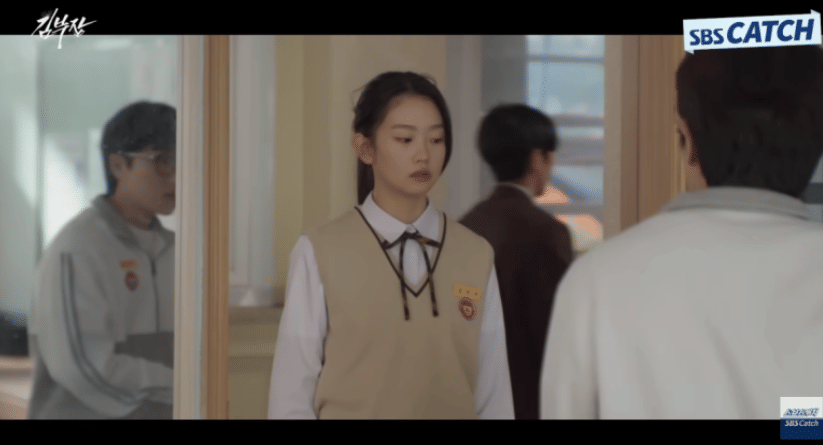 Seorang siswi berseragam sekolah berdiri di koridor sambil berbicara dengan seseorang, dengan latar suasana sekolah dalam drama Agent Kim Reactivated.