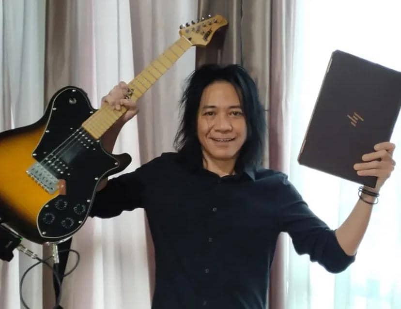 Seorang pria berambut panjang mengenakan kemeja hitam tersenyum sambil memegang gitar listrik dan laptop di depan tirai berwarna netral.