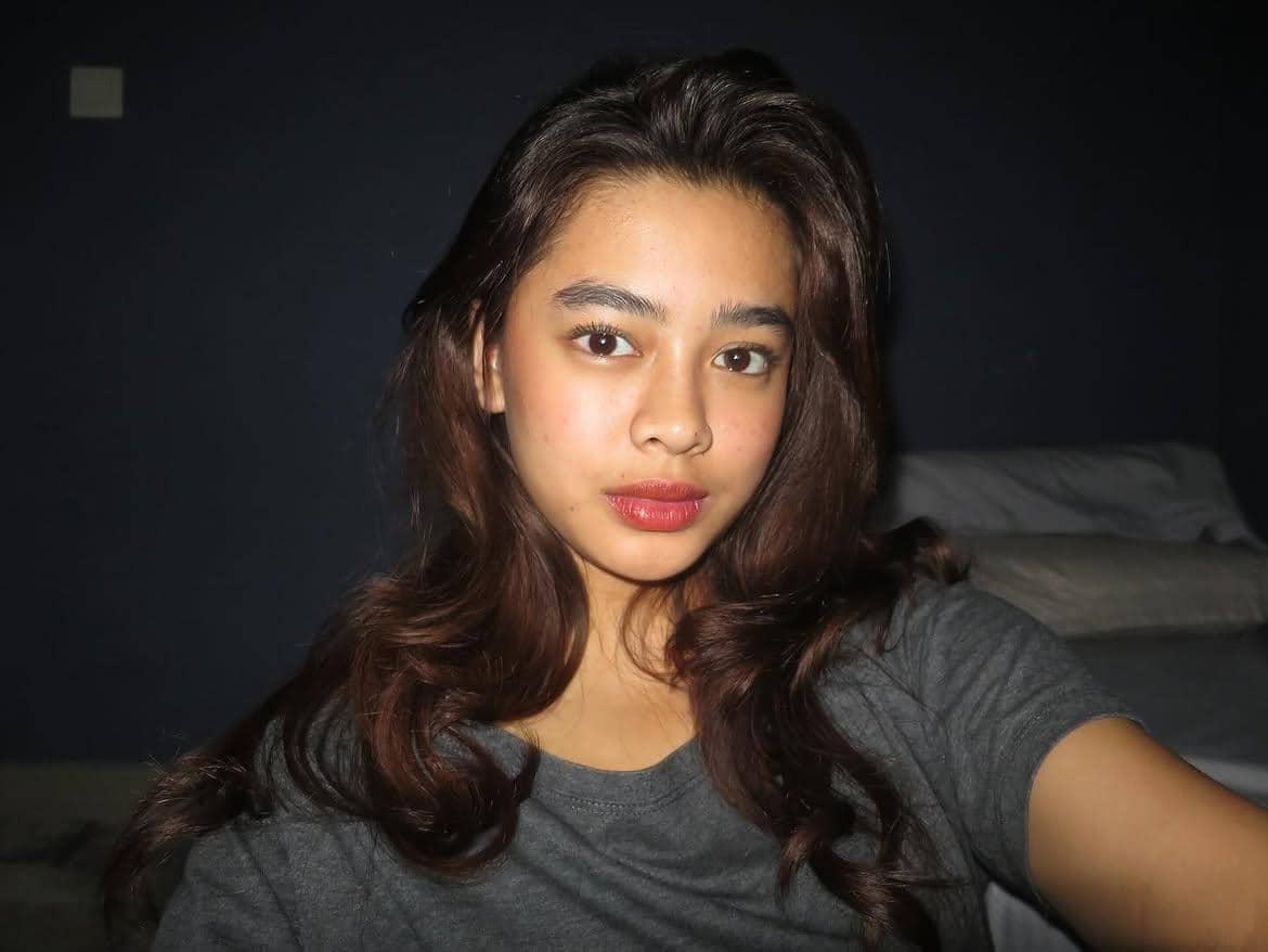 Seorang perempuan berambut panjang bergelombang mengenakan kaus abu-abu berpose di dalam ruangan dengan pencahayaan redup.