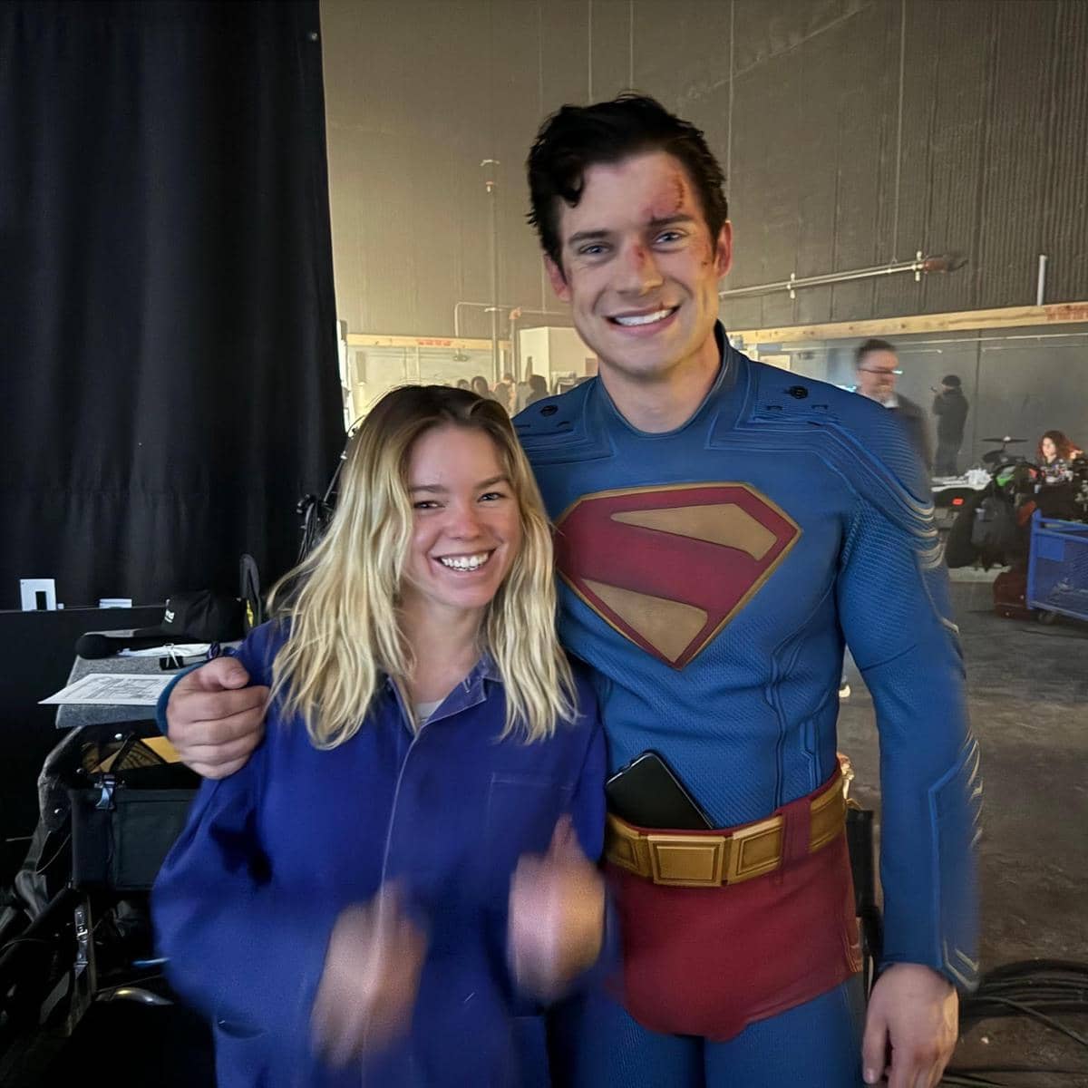 Seorang pria mengenakan kostum Superman berpose sambil tersenyum bersama seorang wanita berambut pirang di lokasi syuting.