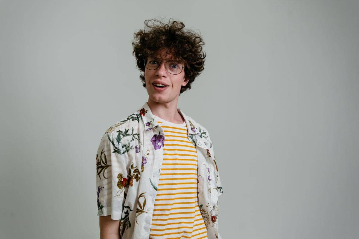 Pria berambut curly top mengenakan kemeja motif bunga di atas kaus bergaris kuning putih dengan latar belakang abu-abu polos.