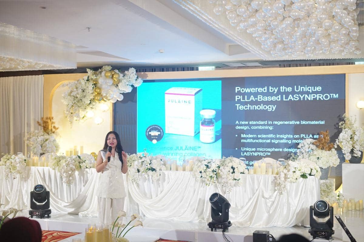 Seorang wanita berbicara di atas panggung dengan latar layar presentasi bertema teknologi kecantikan Julaine of Sweden dan dekorasi bunga putih.