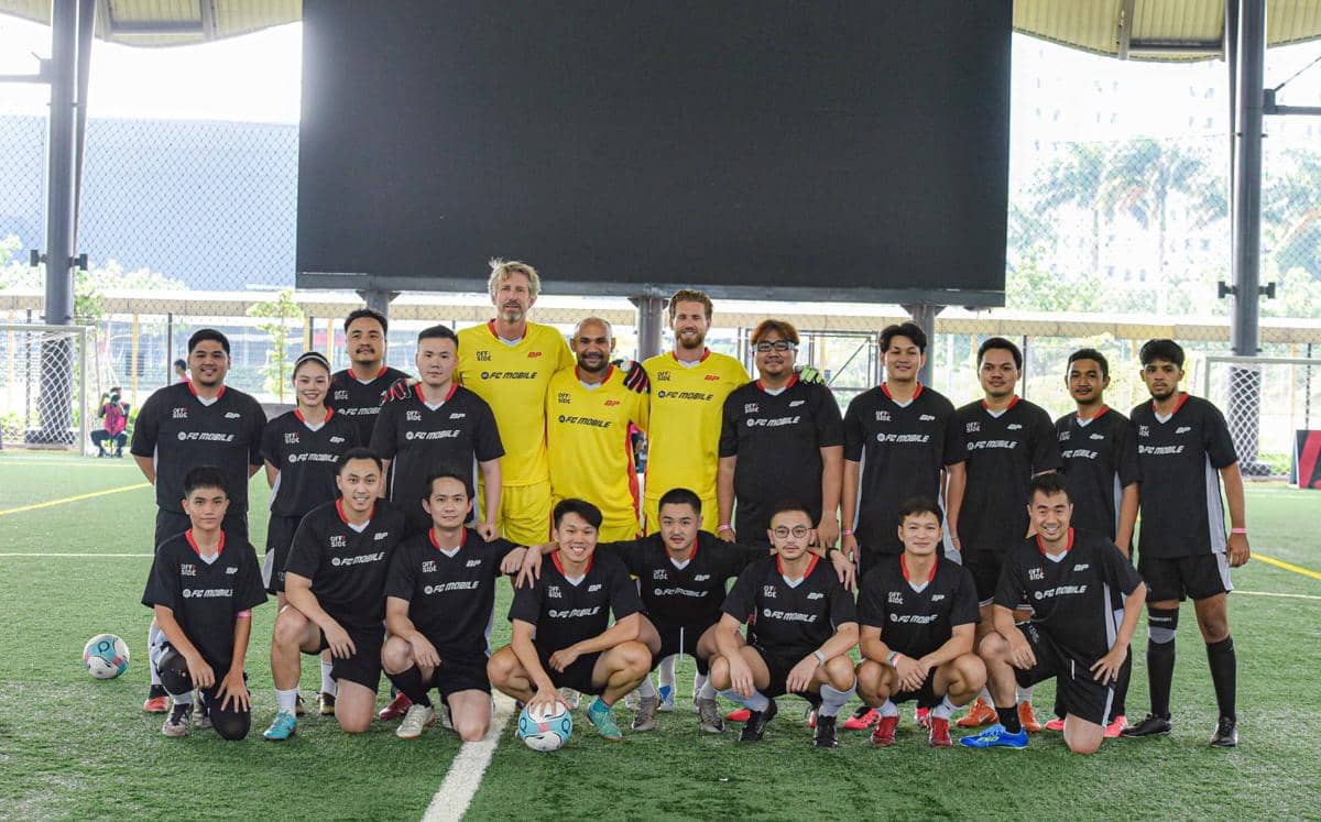 Sekelompok pemain sepak bola berpose bersama di lapangan indoor, mengenakan seragam hitam dan kuning dengan latar gawang dan jaring.