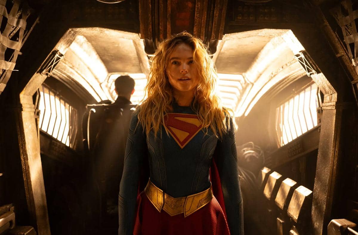 Supergirl berdiri di dalam ruangan logam bercahaya dengan kostum biru dan merah berlogo S di dada, tampak serius menghadapi situasi.