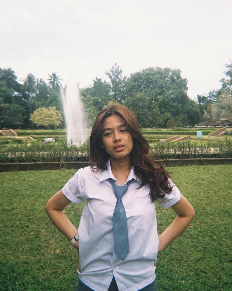 Seorang perempuan mengenakan seragam sekolah putih dan dasi biru berdiri di taman hijau dengan air mancur di latar belakang.
