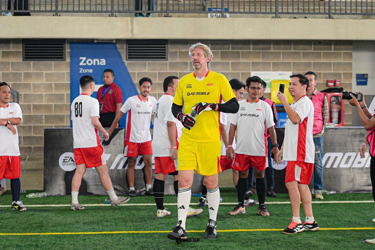 Seorang pria berpostur tinggi mengenakan seragam kiper kuning berdiri di lapangan futsal bersama beberapa pemain lain berseragam putih dan merah.