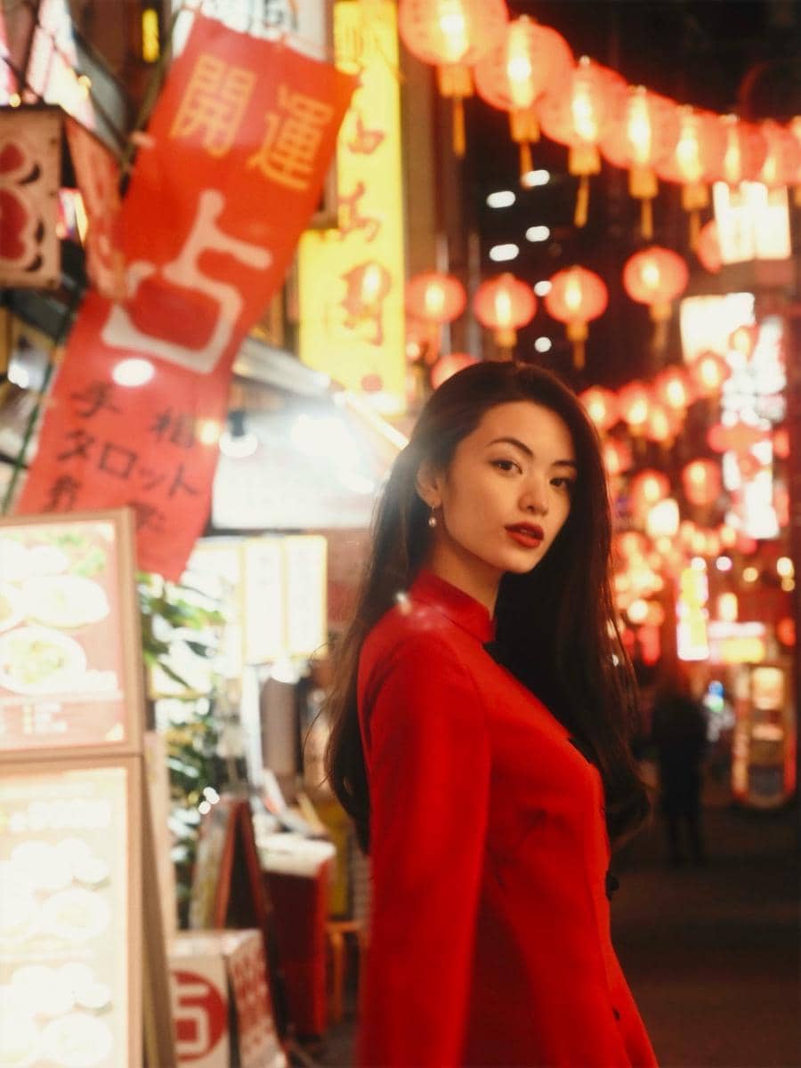 Seorang wanita mengenakan pakaian merah berdiri di jalan kota yang dipenuhi lampion dan papan neon berwarna merah serta kuning di malam hari.