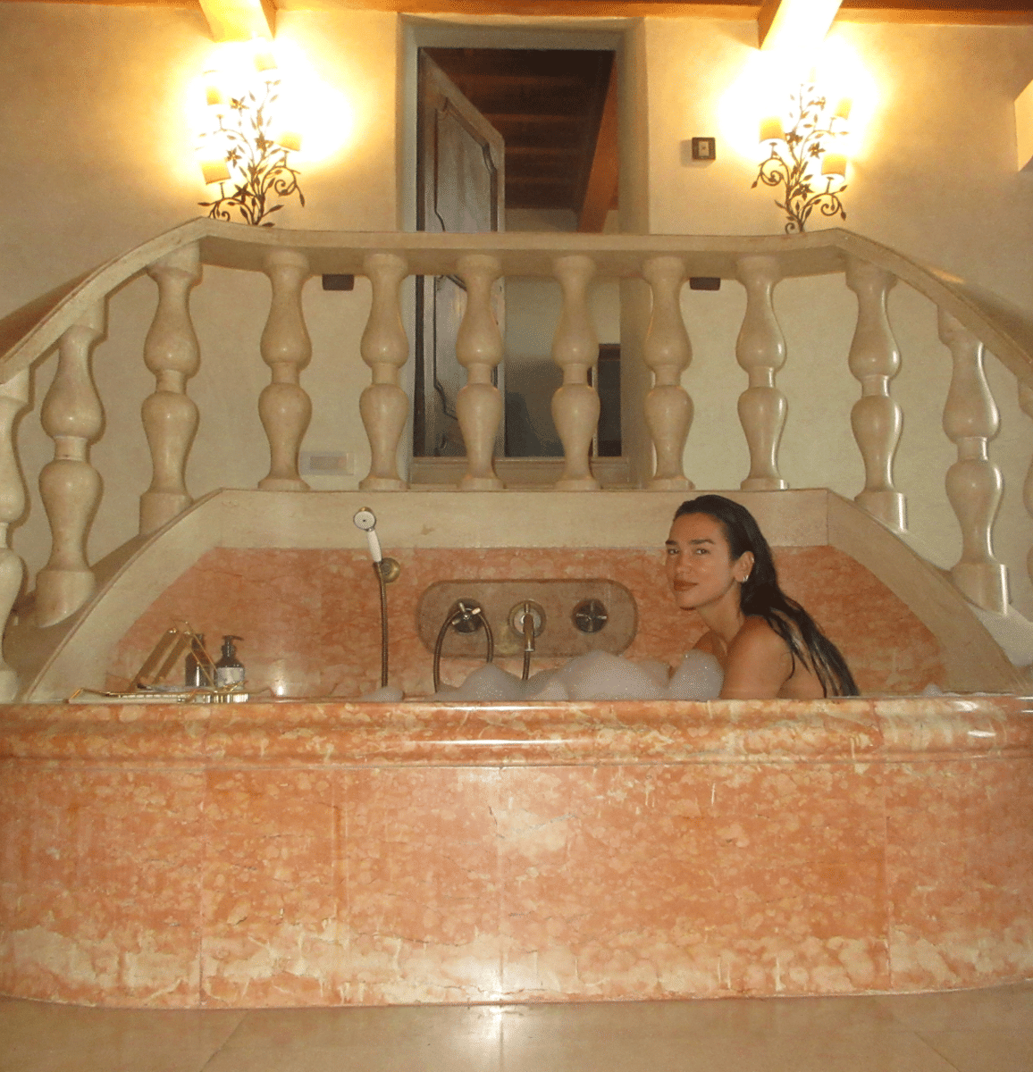 Seorang perempuan berendam di bathtub marmer berwarna oranye muda di kamar mandi bergaya klasik dengan pencahayaan hangat.