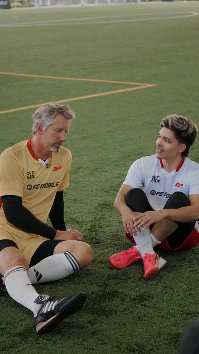 Dua pria duduk di lapangan sepak bola mengenakan seragam olahraga berbeda warna sambil berbincang santai setelah bermain.