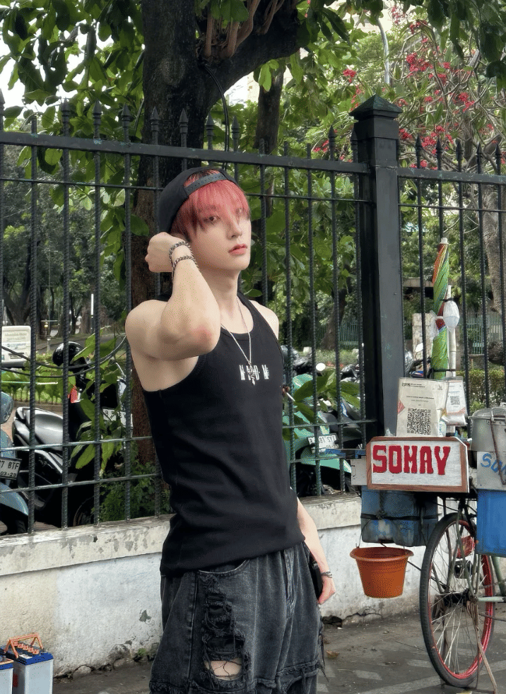 Seorang pria berambut merah mengenakan topi hitam terbalik dan tank top hitam berpose di area trotoar Blok M dengan latar pepohonan dan gerobak es.