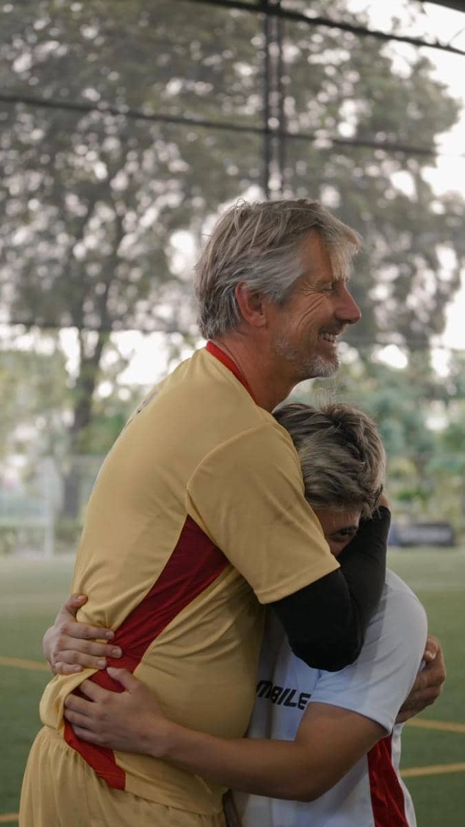 Dua pria berpelukan di lapangan sepak bola, salah satunya mengenakan seragam kuning dan merah, sementara yang lain berbaju putih.