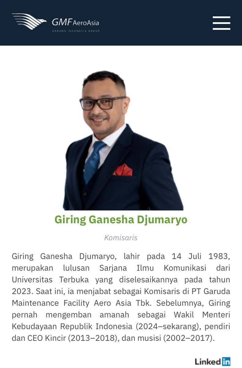 Seorang pria mengenakan setelan jas biru tua dengan dasi biru dan saputangan merah berdiri tersenyum di latar putih dengan logo GMF AeroAsia di atasnya.