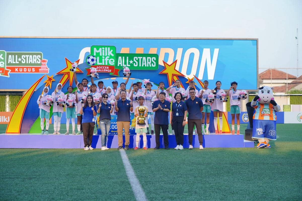 Tim sepak bola muda berpose di podium juara bersama panitia dan maskot setelah memenangkan turnamen Milk Life Soccer Challenge All Stars 2026.