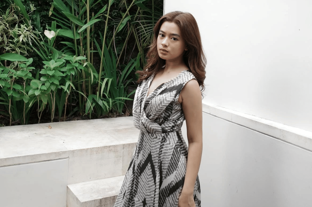 Seorang perempuan berambut panjang mengenakan gaun bermotif hitam putih berdiri di dekat dinding putih dengan latar tanaman hijau.