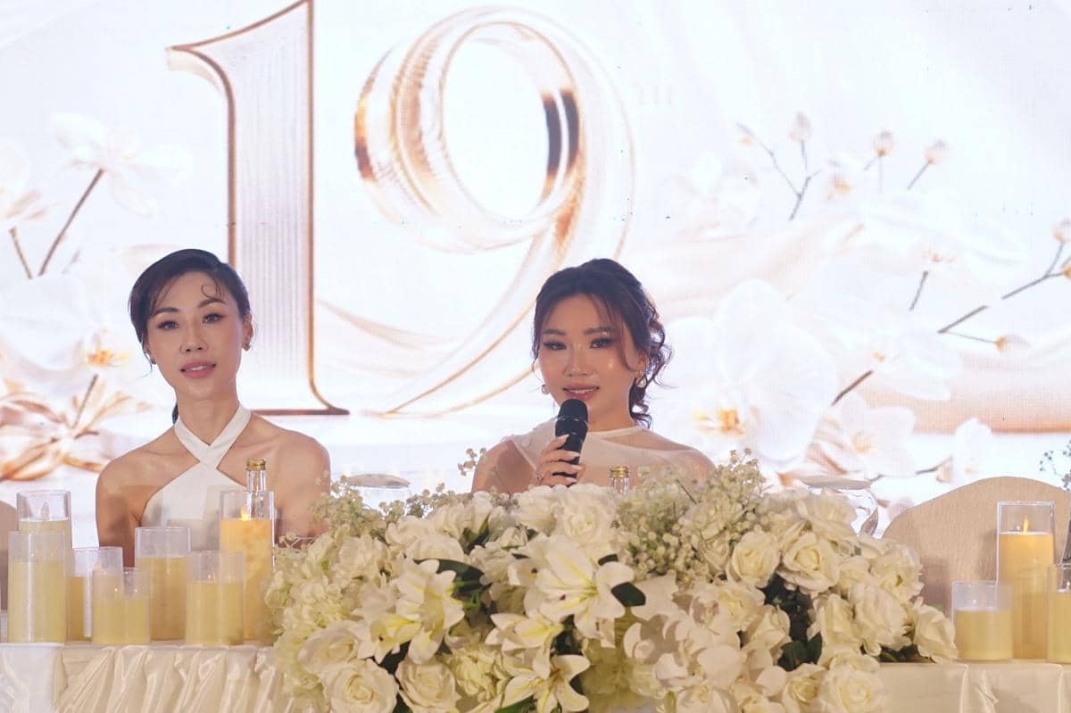 Dua wanita berbusana putih duduk di meja berhias bunga dan lilin, salah satunya memegang mikrofon di acara bertema elegan.