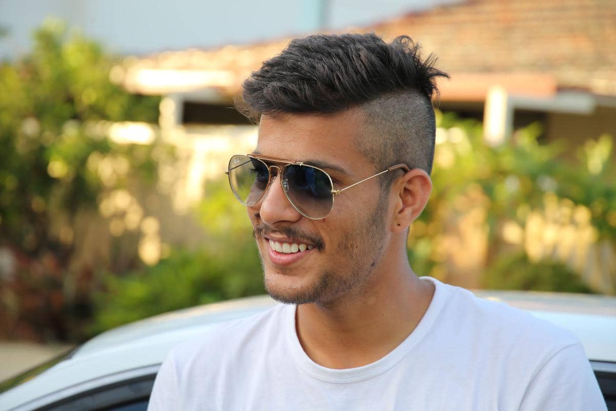 Pria muda mengenakan kacamata hitam dengan gaya rambut front spikes dan sisi kepala dicukur pendek di luar ruangan.