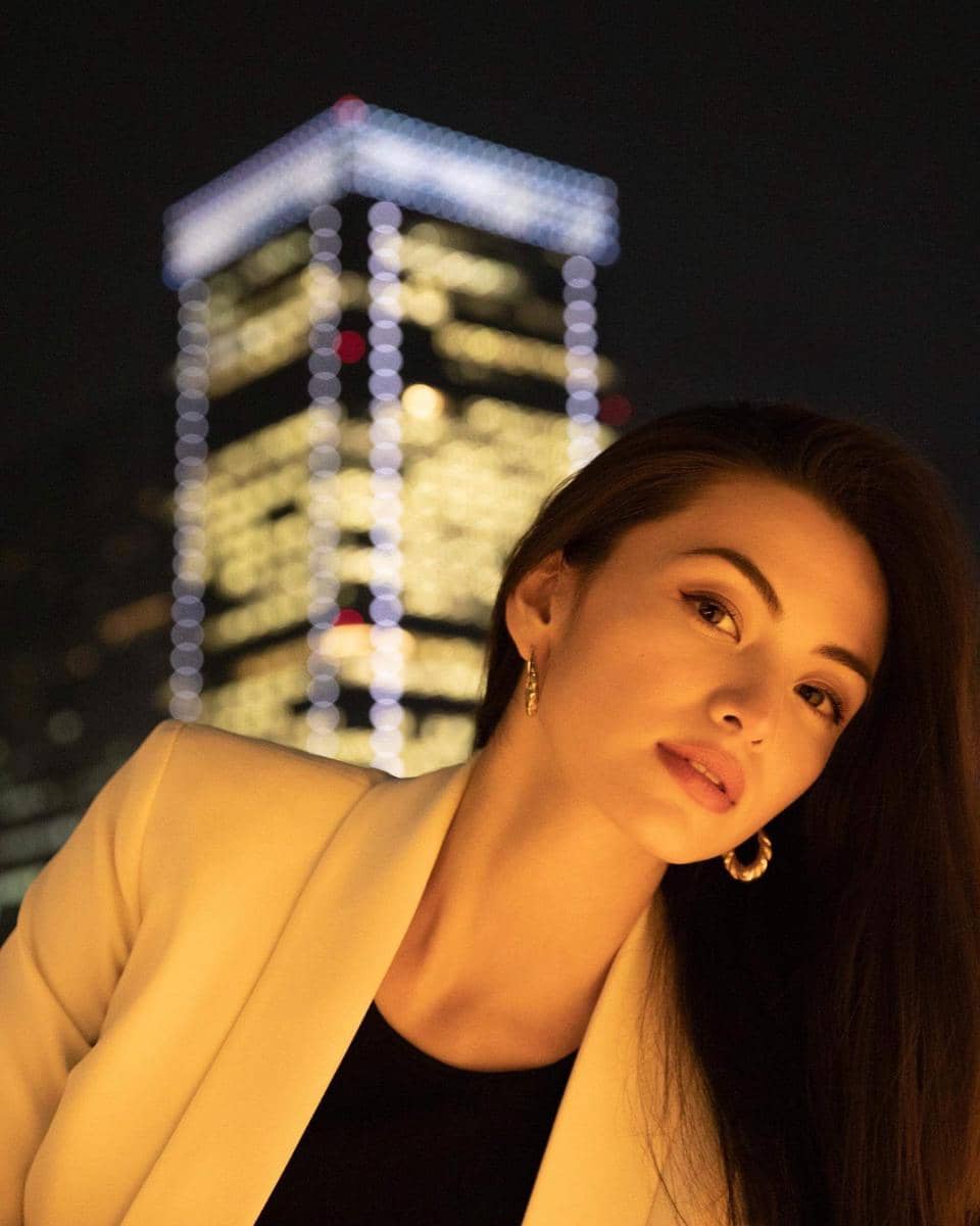 Seorang wanita berambut panjang mengenakan blazer krem berpose di malam hari dengan latar gedung tinggi bercahaya di belakangnya.