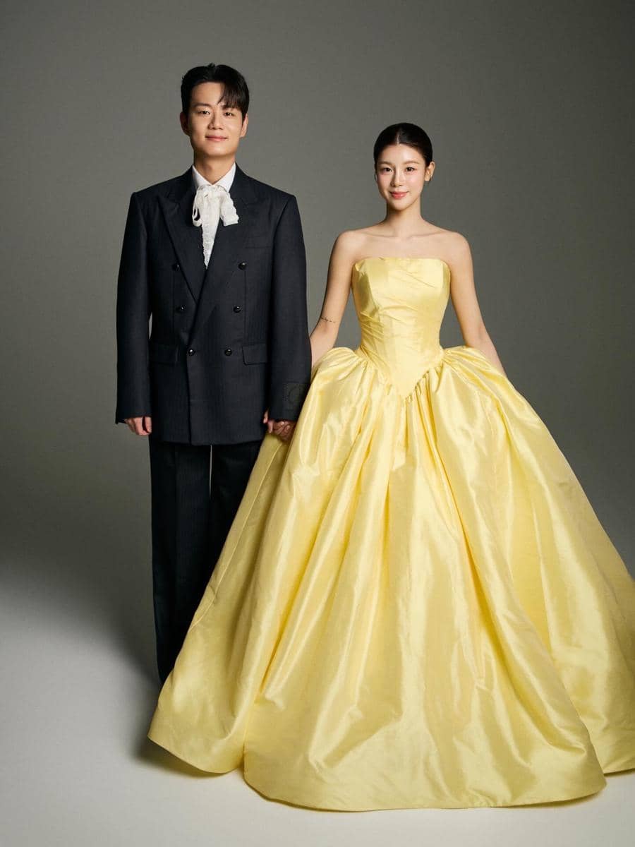 Sepasang pria dan wanita berdiri berdampingan mengenakan busana formal, pria dengan setelan hitam dan wanita dengan gaun kuning elegan.