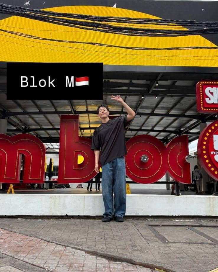 Seorang pria berpose sambil mengangkat tangan di depan tulisan besar Blok M di area publik Jakarta dengan latar warna kuning dan merah.