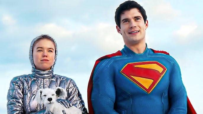 Supergirl mengenakan pakaian perak bersama Superman berseragam biru dan merah, dengan anjing putih Krypto di pelukannya di bawah langit cerah.