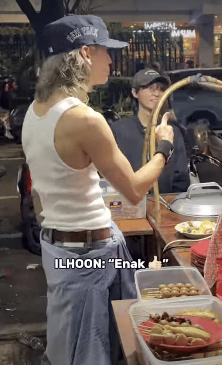 Seorang pria mengenakan topi bertuliskan New York dan tank top putih sedang membeli makanan kaki lima di kawasan Blok M pada malam hari.